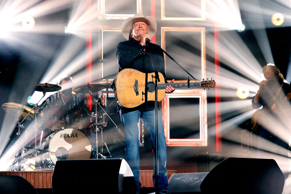 Alan Jackson performing on stage under bright lights, singing into the microphone while holding his acoustic guitar.
