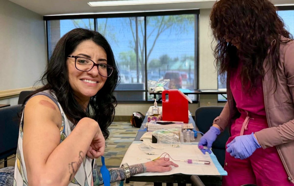 A smiling participant seated at a table has a blood sample drawn by a gloved phlebotomist at a CMT Biobank collection event, with labeled collection tubes and supplies laid out on the table.