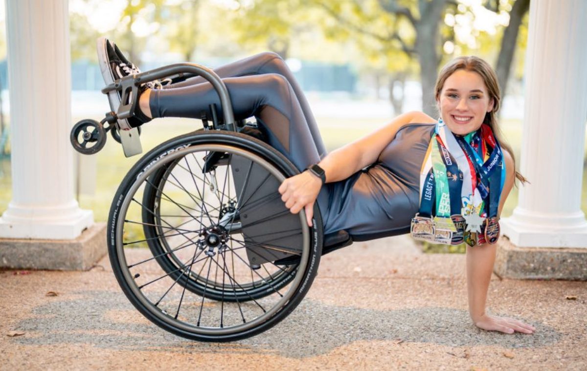 A young woman in a patterned jacket sitting on the ground beside her racing wheelchair, outdoors between white stone columns.