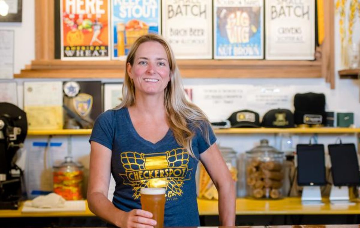 A woman in a Checkerspot Brewing T-shirt holding a glass of beer behind the taproom counter, with framed beer labels on the wall behind her.