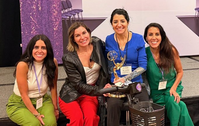 Four women together on stage at a CMT Summit, one seated in a power wheelchair holding an award.