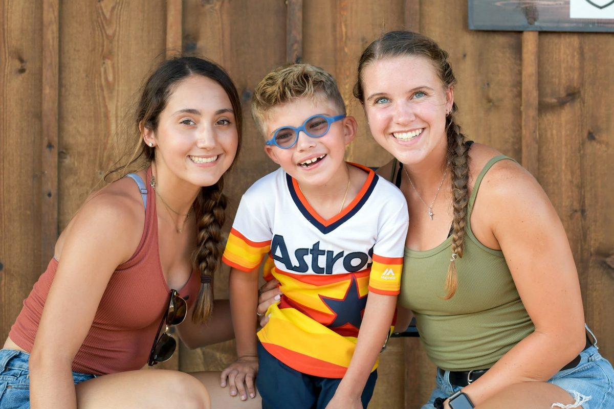 Jaxson, a young boy in a baseball jersey and glasses, smiling between two young women in front of a wooden wall.