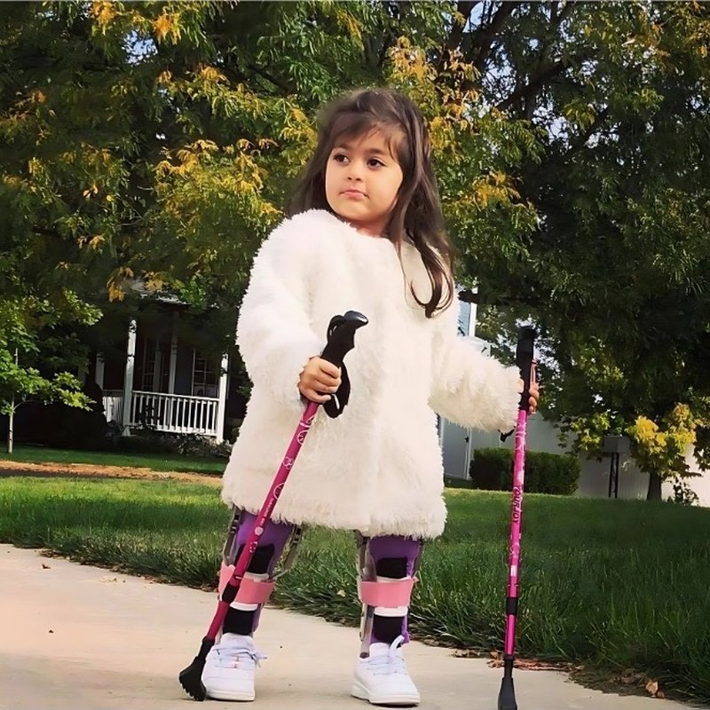 A young girl in leg braces walking outdoors with two pink walking poles.