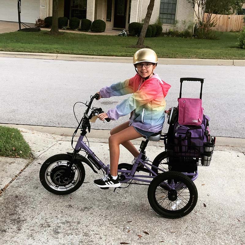 Addie riding her adaptive tricycle, wearing a helmet, on a sunny street.