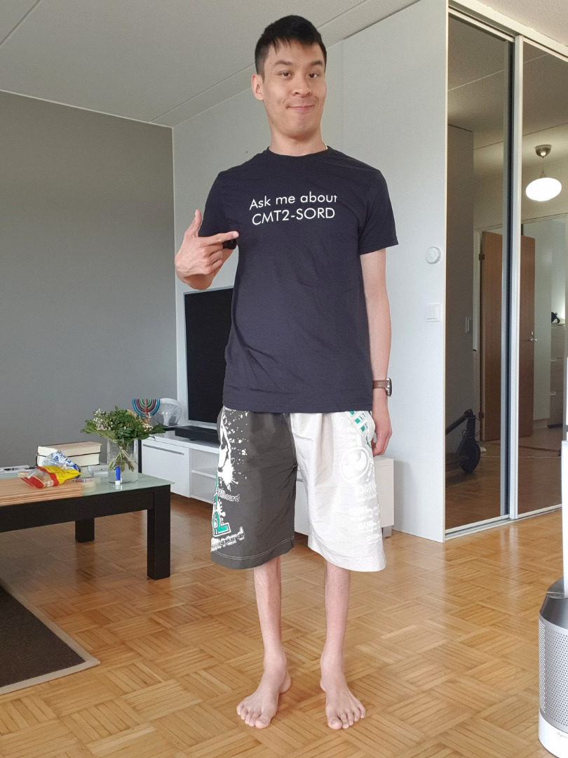 Christopher Gullmans standing barefoot in his living room, pointing at his navy t-shirt printed with the words Ask me about CMT2-SORD.