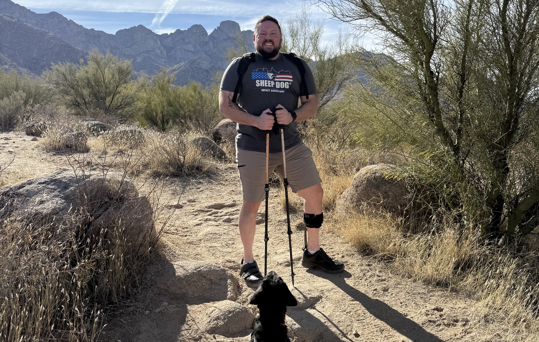 Daniel Blevins standing on a desert trail in Arizona, leaning on trekking poles and wearing an ankle-foot orthosis, with his black service dog sitting on the path in front of him.