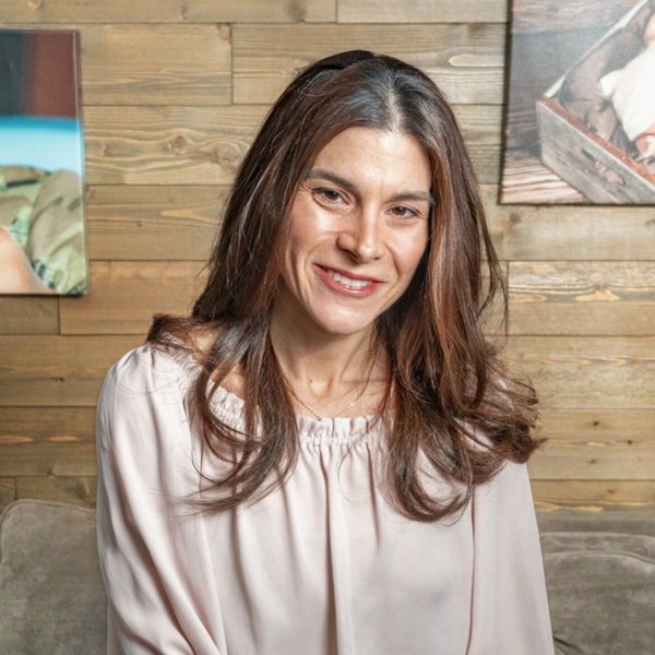 Jennifer DeSetto smiling, seated in front of a wood plank wall hung with framed photographs.