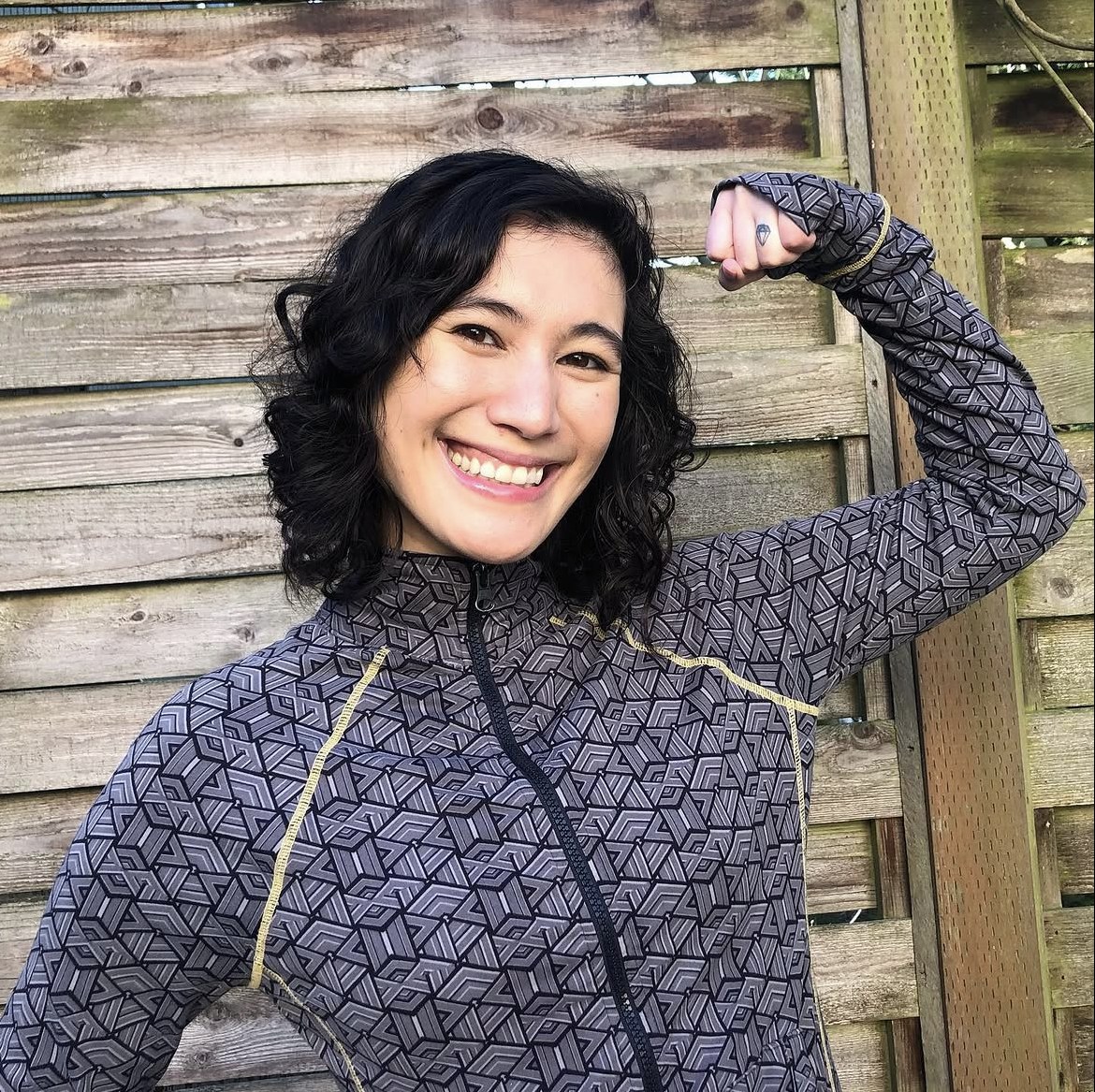 Julie Stone smiling and flexing one arm in front of a wooden fence, wearing a patterned training jacket.