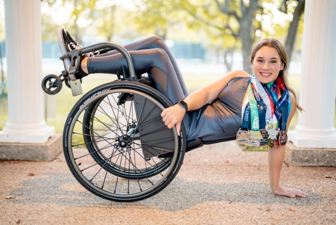 Skyler Fisher reclining against her racing wheelchair on a paved walkway between white columns, wearing a stack of triathlon medals around her neck.