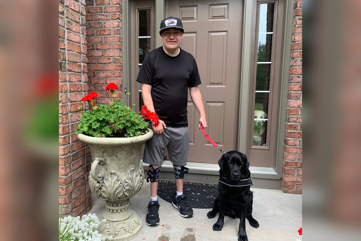 Zach standing at his front door with his black service dog, wearing leg braces.