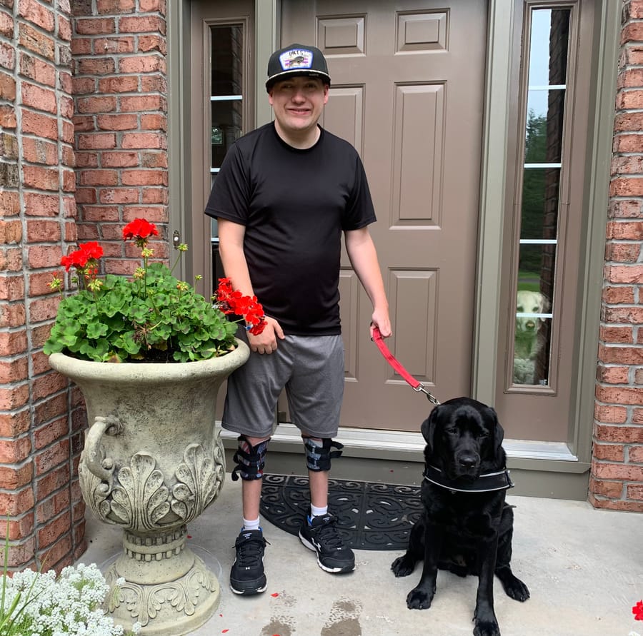 Zach standing at his front door with his black service dog, wearing leg braces.