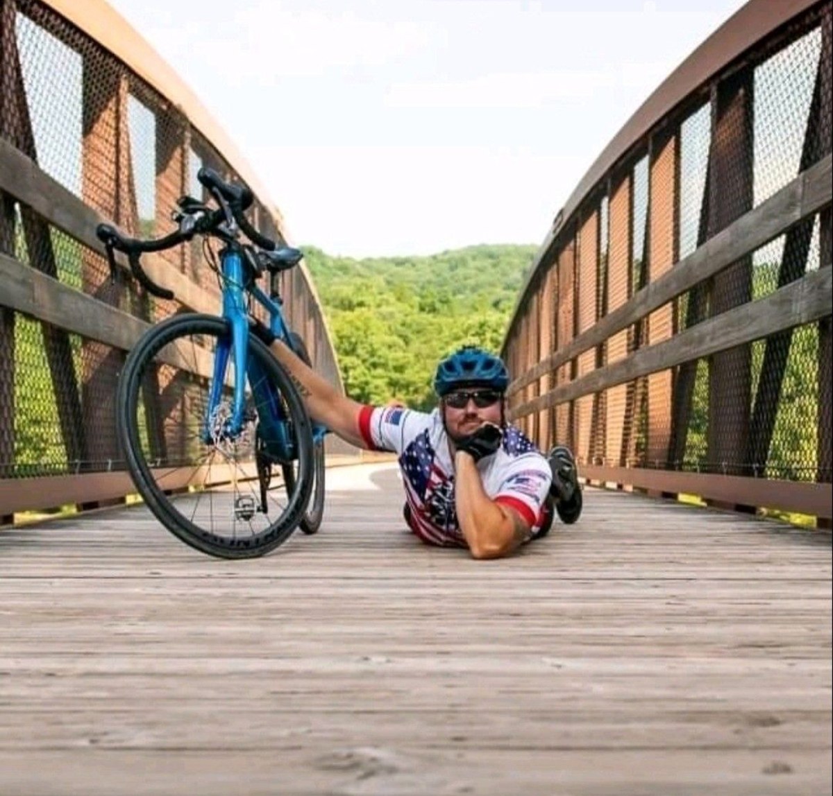 Daniel Blevins in cycling kit lying on a wooden bridge behind his road bike, which stands upright in the frame.