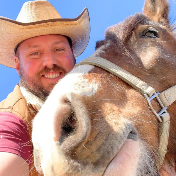 Daniel Blevins in a straw cowboy hat, cheek to cheek with a horse in bright sunshine.