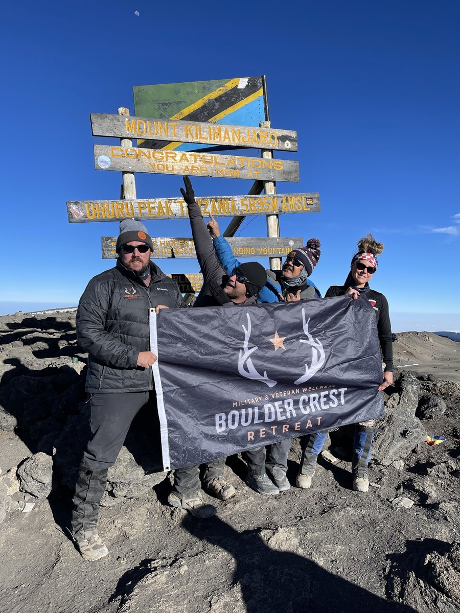 Daniel Blevins, at left, with his team at the Uhuru Peak sign on the summit of Mount Kilimanjaro, holding a Boulder Crest Retreat military and veteran wellness banner.