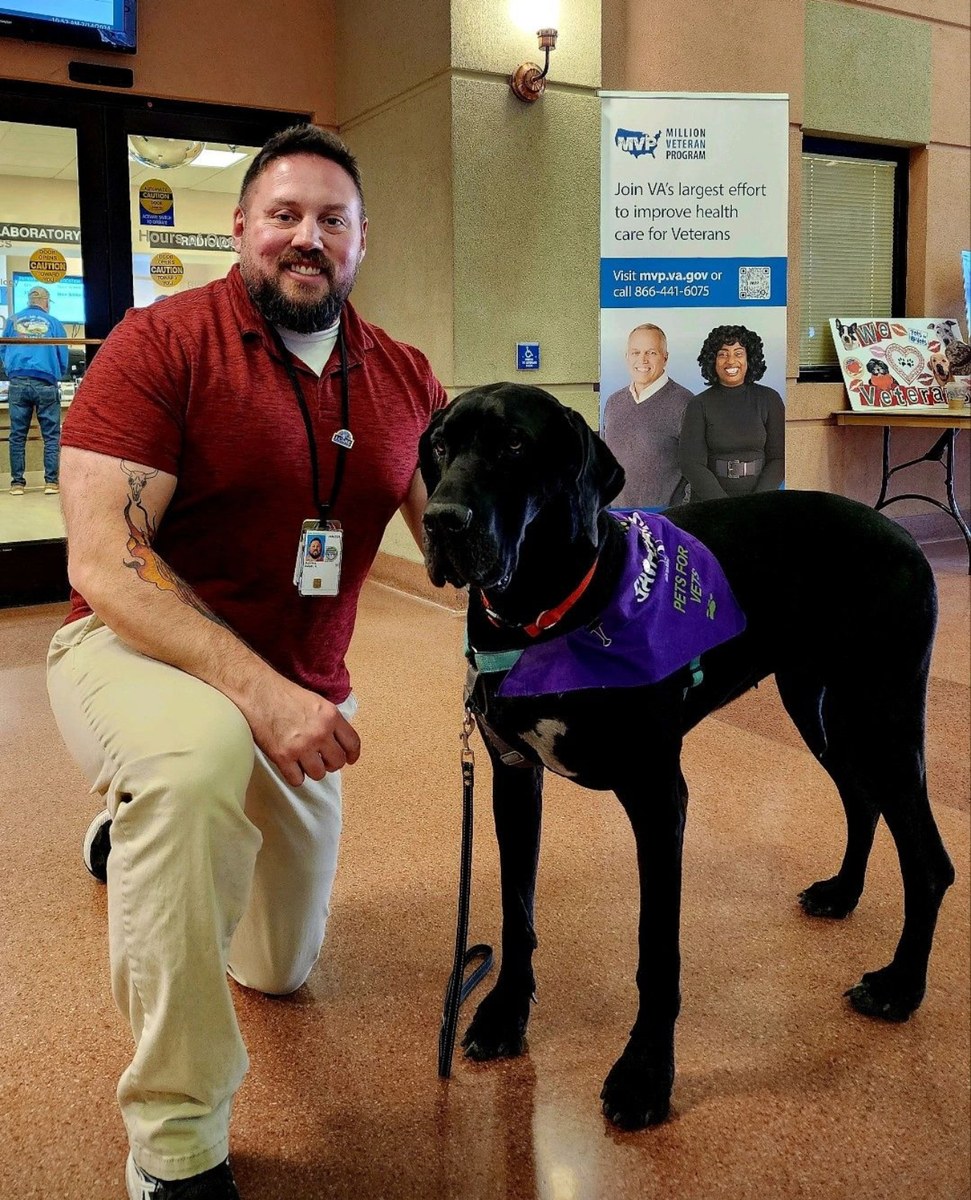 Daniel Blevins kneeling beside his black service dog, who is wearing a purple service vest, at a VA medical centre.