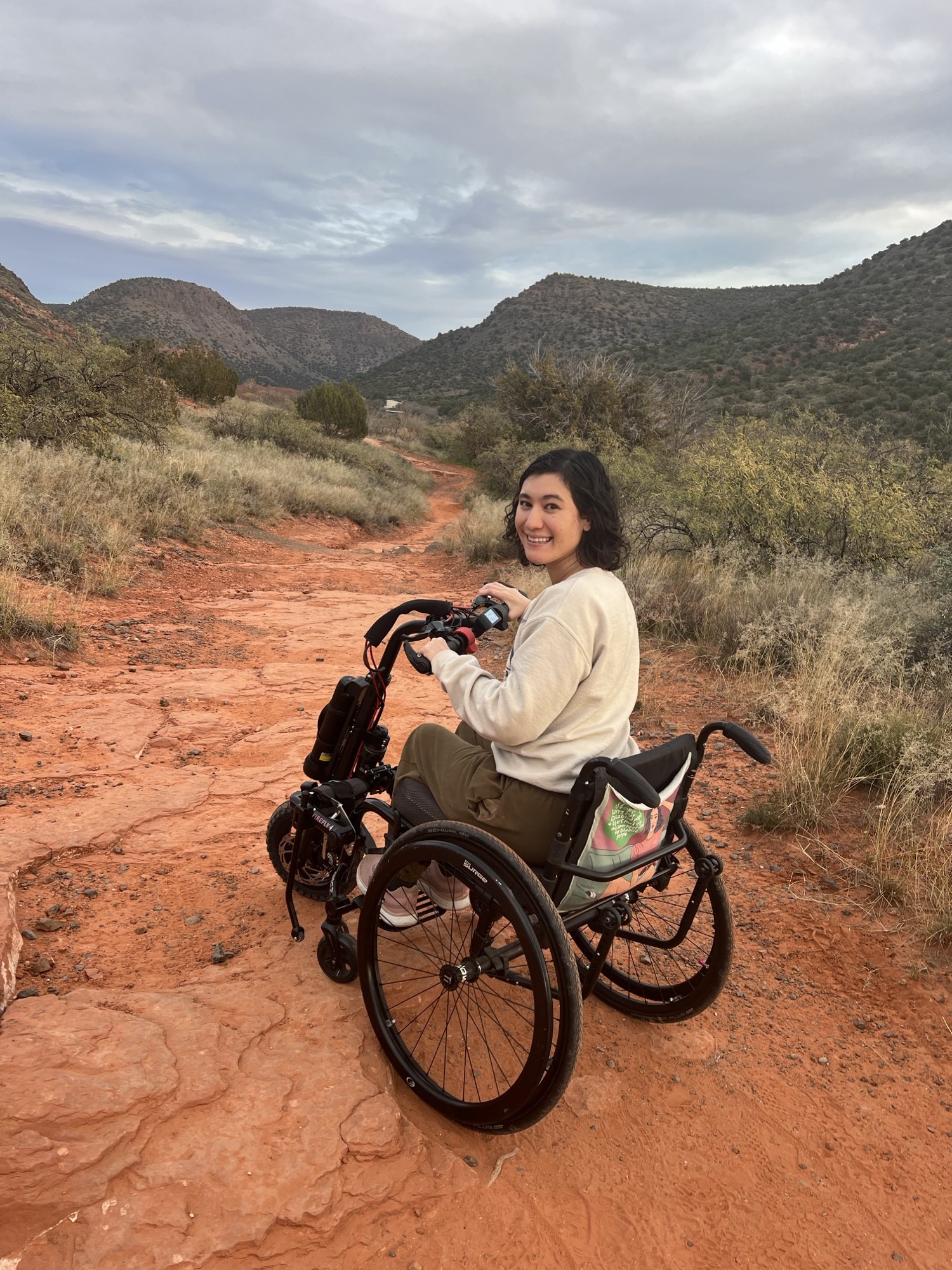 Julie Stone smiling on a red rock trail in her wheelchair with a Rio Firefly power attachment on the front.