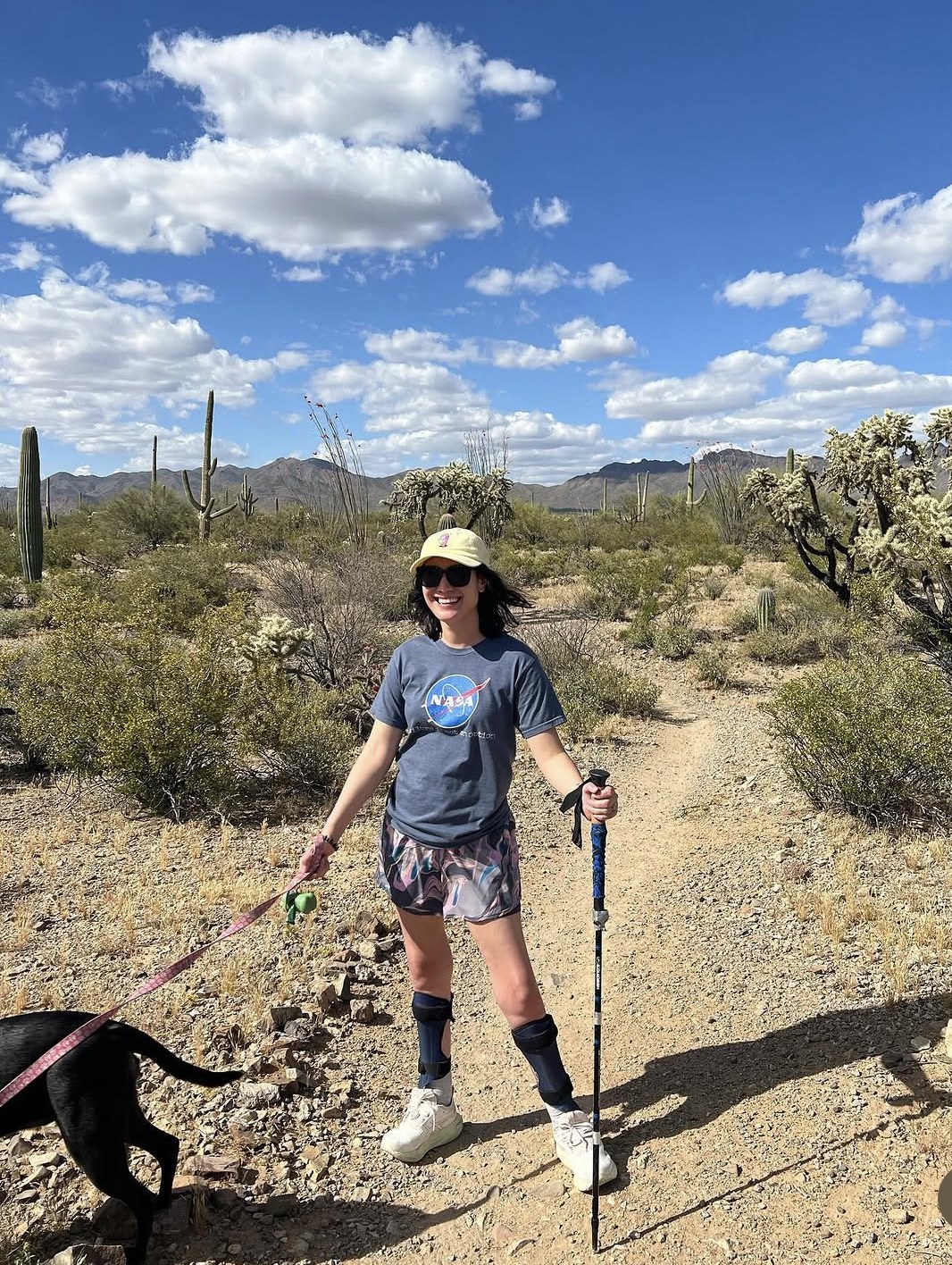 Julie Stone on a desert trail among saguaro cactus, walking her dog in ankle-foot orthoses with a trekking pole.