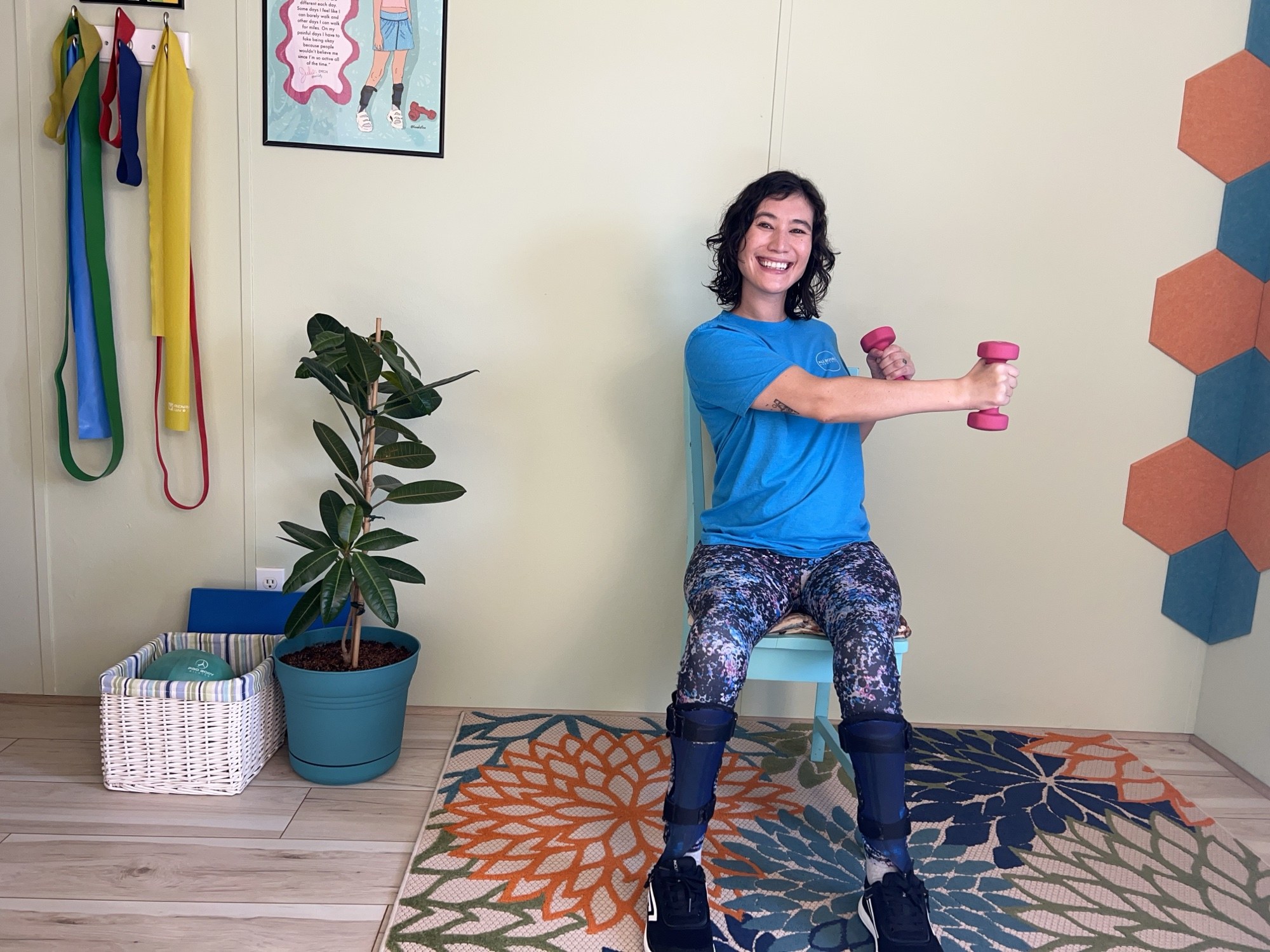 Julie Stone training with pink dumbbells in her studio, wearing her leg braces, resistance bands hanging on the wall behind her.