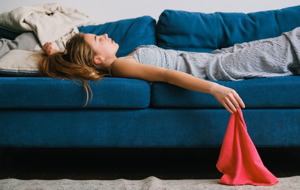 A woman lying back on a blue couch with her head resting on the cushions and one arm draped over the side, a pink cloth in her hand.