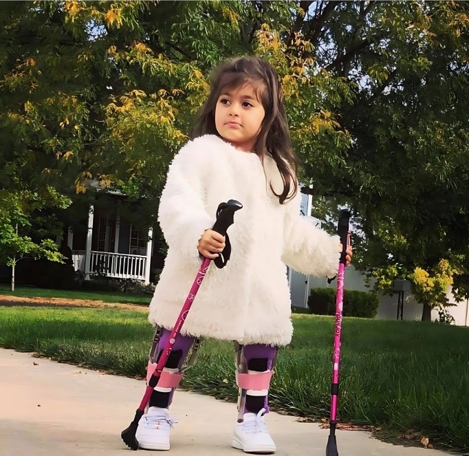 A young girl in a fluffy white jacket walking outdoors on a sidewalk using two pink walking poles, wearing patterned leg braces over white boots.