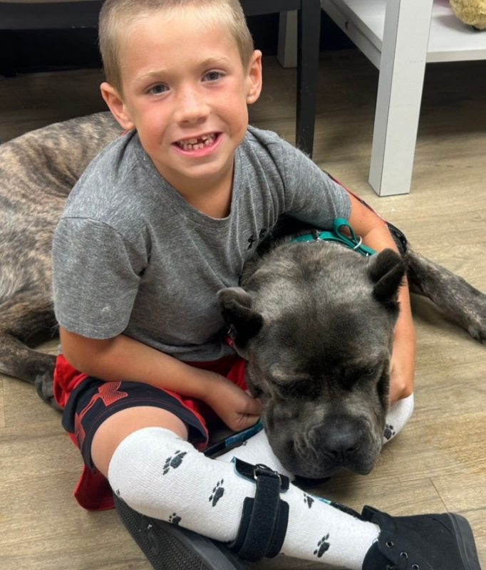 A smiling young boy sits on the floor with his arm around a large, calm brindle dog, wearing paw-print socks and an ankle-foot orthosis brace.