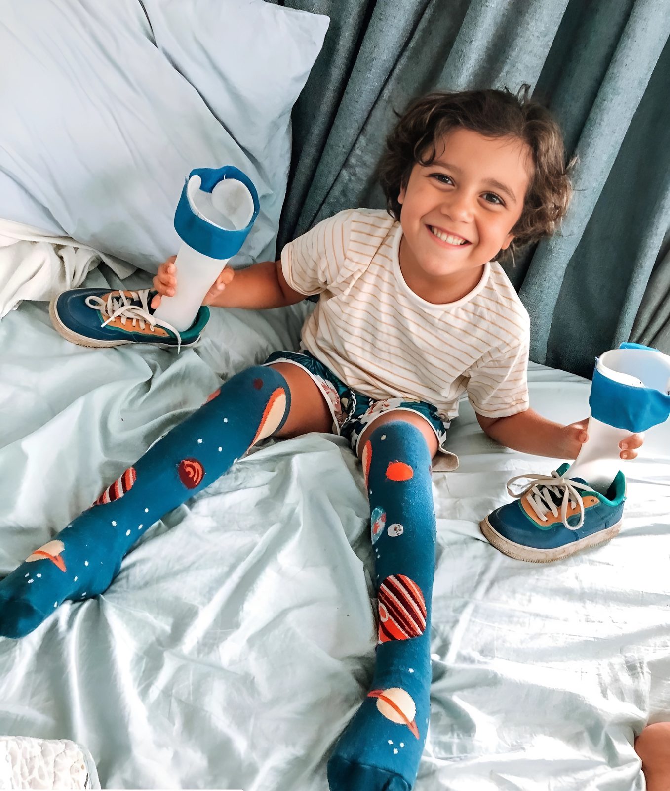 A smiling young boy in planet-patterned knee socks sits on a bed holding his ankle-foot braces, his sneakers beside him.