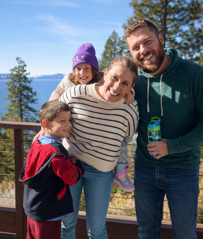A smiling family of four stands close together on an outdoor deck overlooking a lake and pine trees, two parents with their young son and daughter.