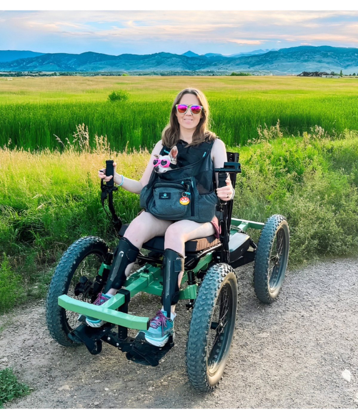 A woman in sunglasses riding an all-terrain outdoor wheelchair on a gravel path through green fields.