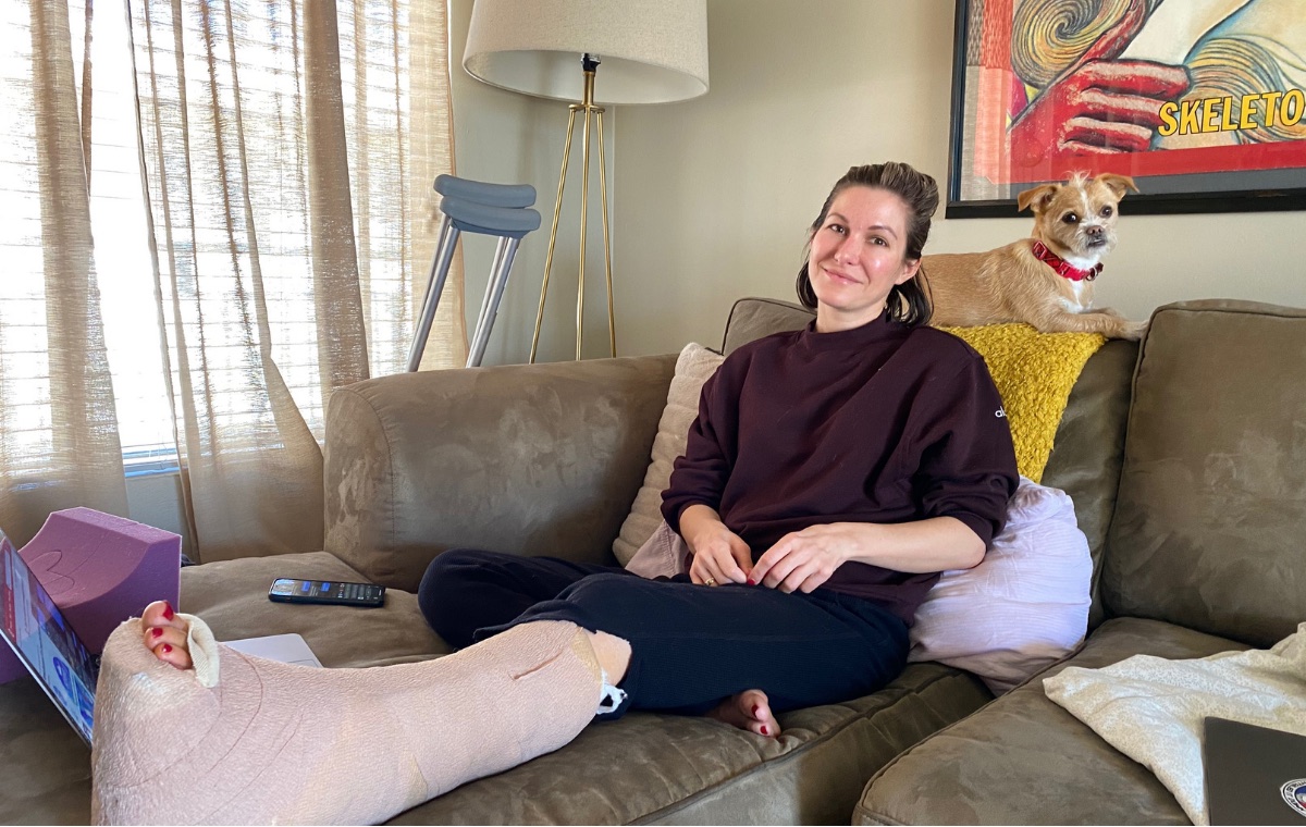 A smiling woman relaxing on a couch at home with a small dog beside her, one leg resting on a cushion.