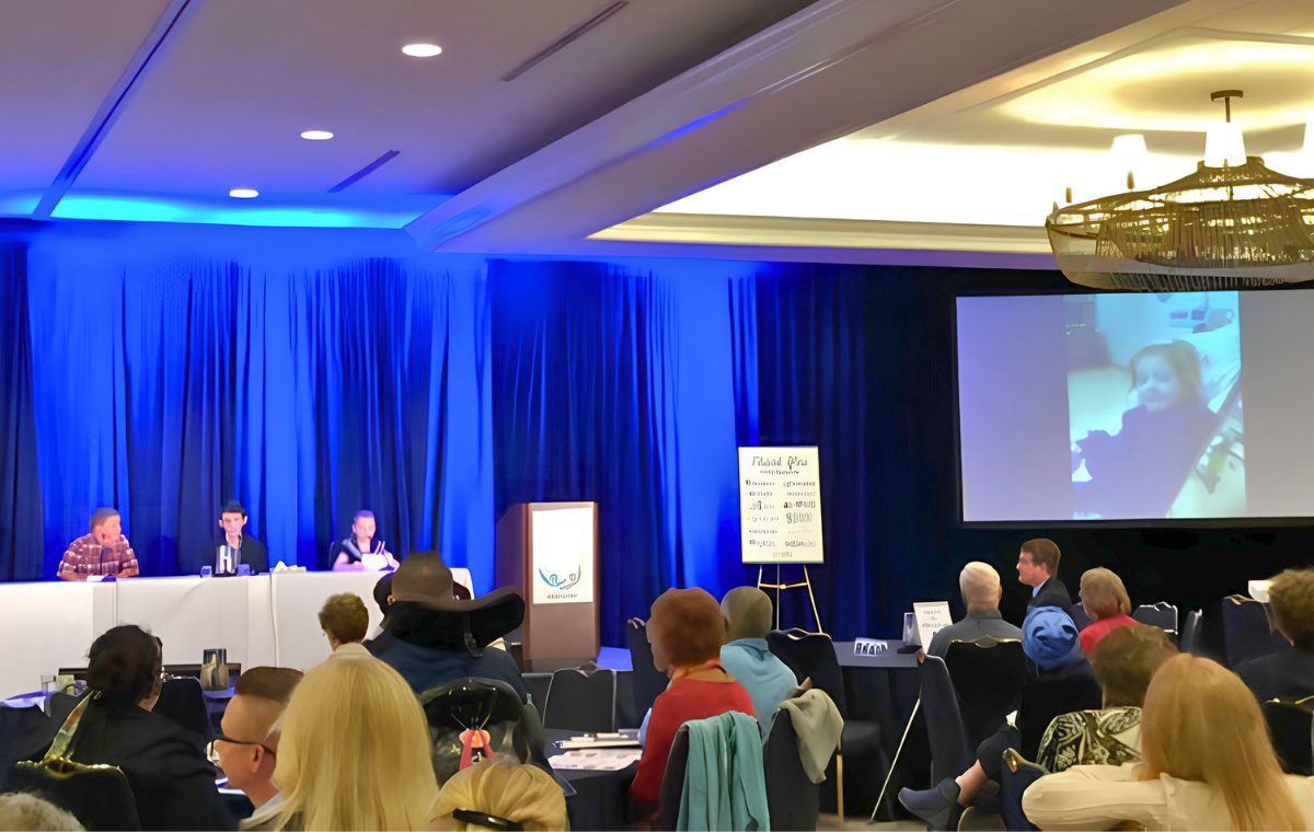 A PFDD meeting in a hotel ballroom with blue lighting, showing a panel seated at a head table, a seated audience, and a remote participant on a large projection screen.