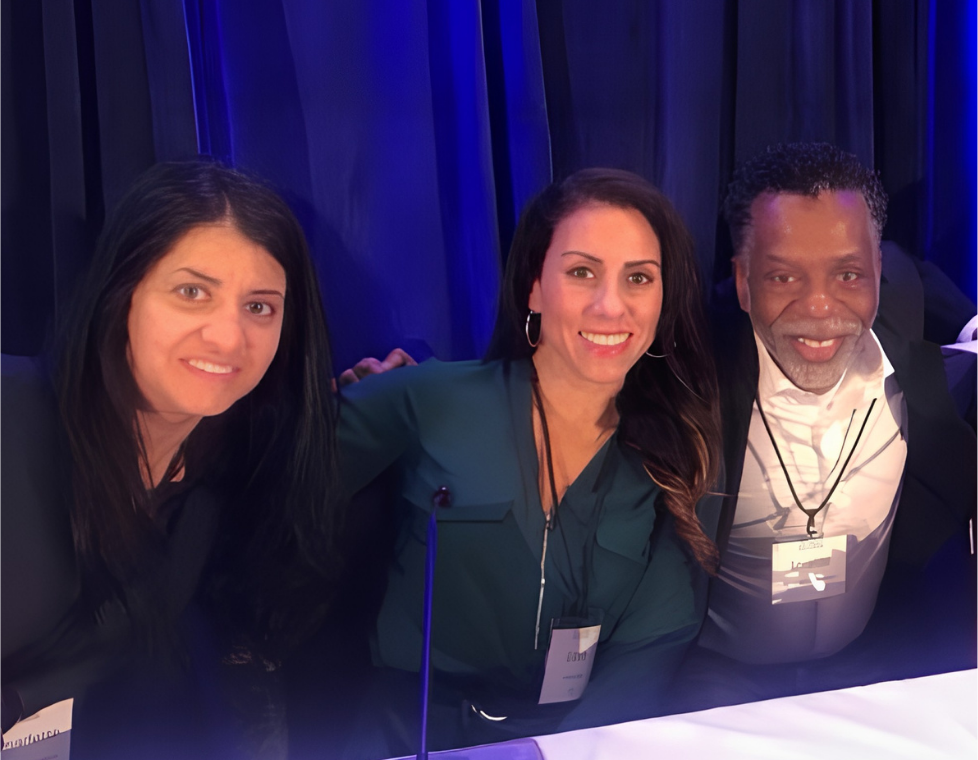 Three CMT community members seated together at a panel table in front of blue draping at the PFDD meeting, wearing name badges and smiling.