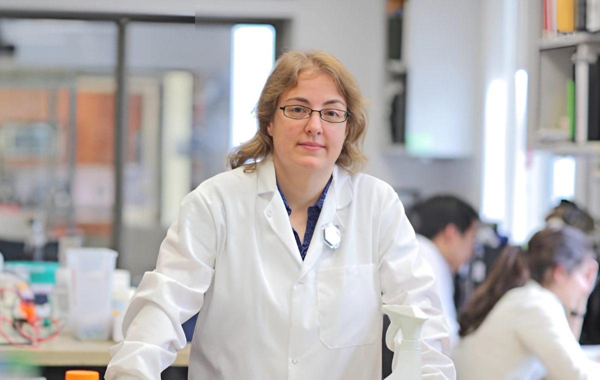 A researcher in a white lab coat stands at a laboratory bench surrounded by equipment and reagents, with colleagues working in the background.
