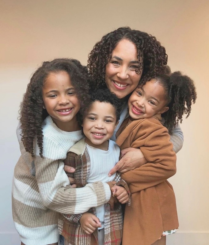 A smiling mother with her arms around her three young children, all leaning in close together for a warm family portrait.