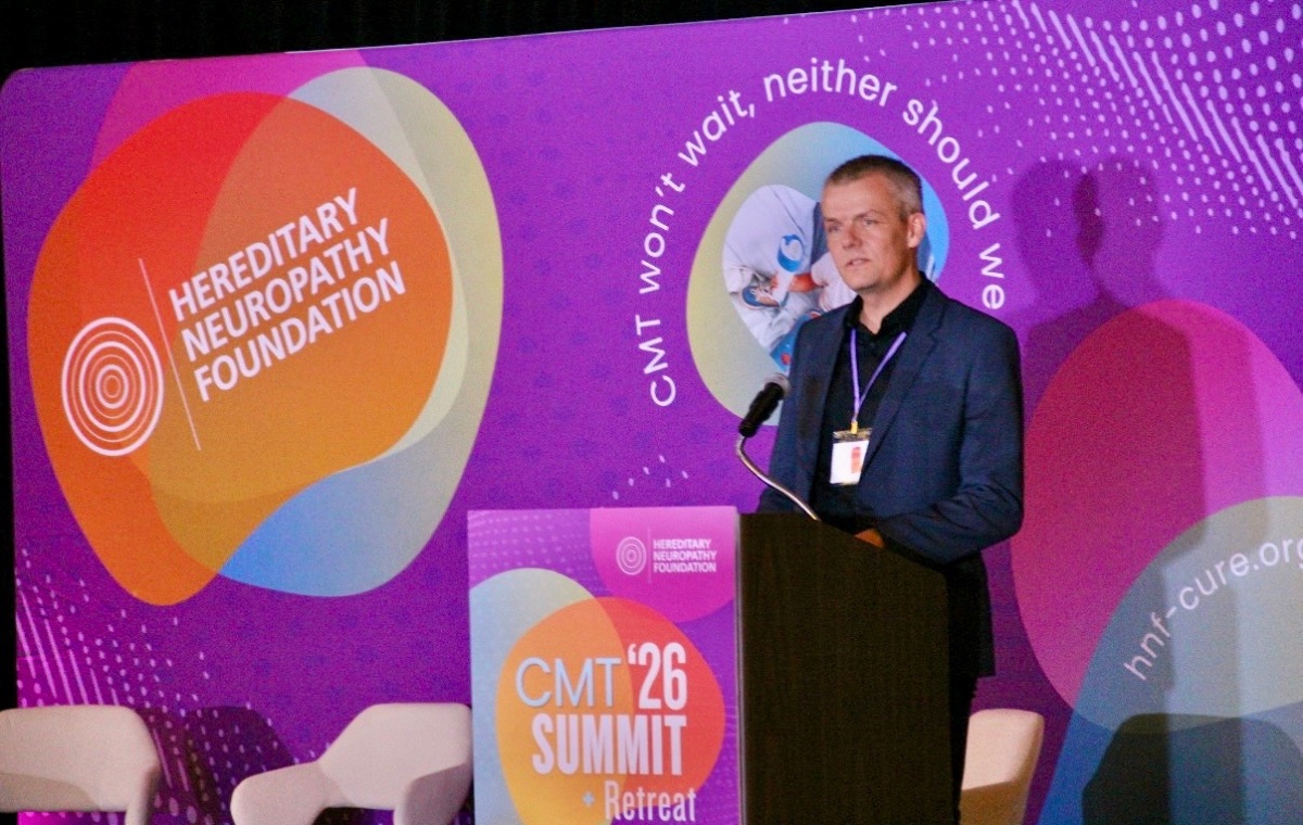 A speaker presenting at a podium on the CMT Summit stage, in front of a Hereditary Neuropathy Foundation backdrop.