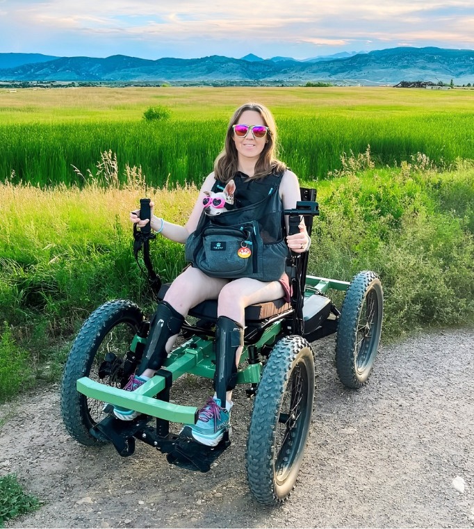 A woman in an all-terrain off-road wheelchair on a gravel path, wearing leg braces and sunglasses, with green fields and mountains behind her.