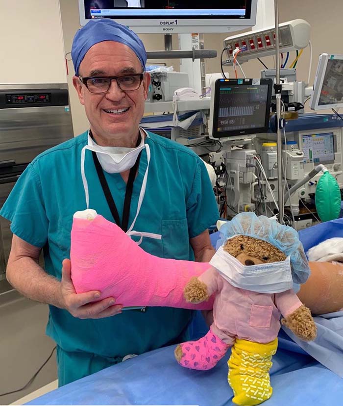 Dr. Glenn Pfeffer in surgical scrubs in an operating room, holding a child’s leg in a pink cast beside a teddy bear wearing a matching cast and surgical cap.
