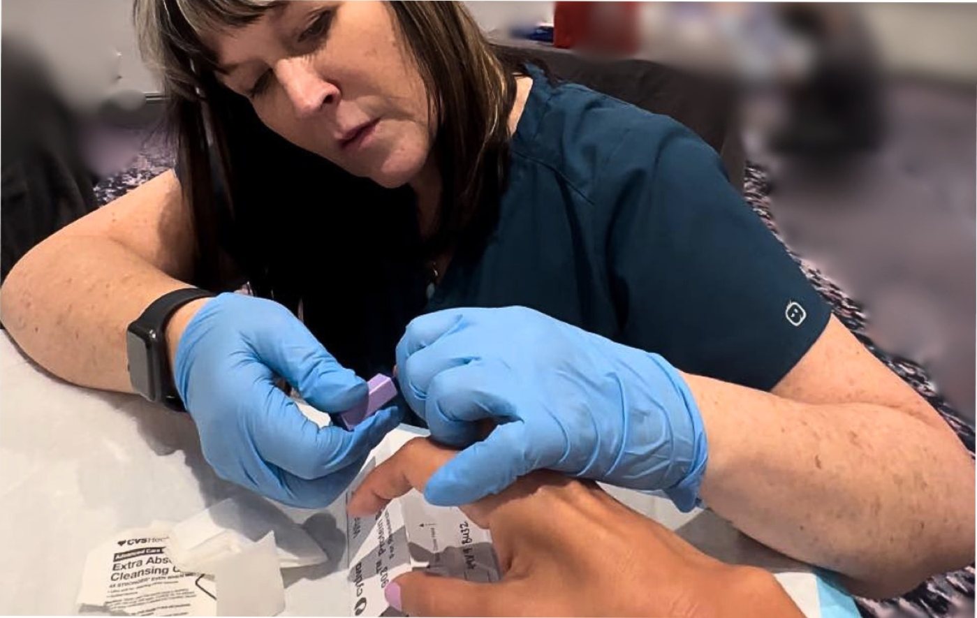 A researcher in gloves collecting a fingerstick blood sample from a participant at an onsite research station.