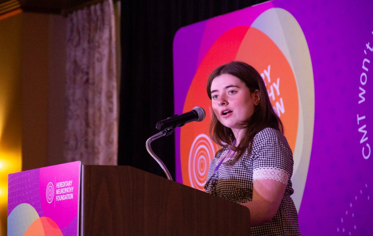 A young woman speaks at a wooden podium bearing the Hereditary Neuropathy Foundation logo, in front of a purple stage backdrop reading CMT Won't Win, at an HNF summit.
