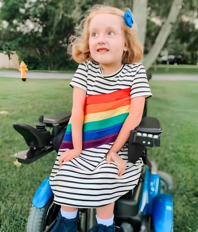 A smiling young girl in a rainbow dress sits on her power wheelchair in a sunny park.