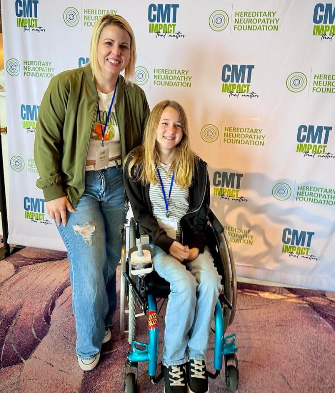 A woman stands beside a smiling young girl seated in a wheelchair, both wearing attendee badges, in front of a Hereditary Neuropathy Foundation and CMT Impact backdrop at a patient-focused CMT event.