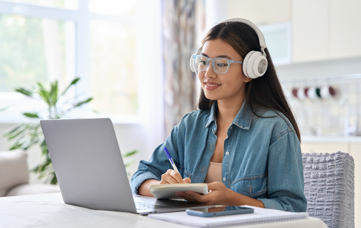 A smiling young woman wearing headphones and glasses attends a webinar from home, taking notes in a notebook while looking at her laptop at a sunlit kitchen table.