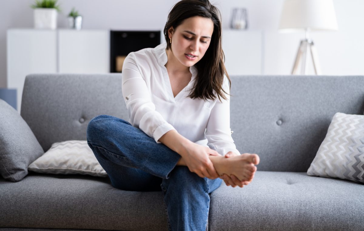 A woman sits on a sofa at home holding her bare foot, reflecting the foot and lower-leg symptoms that often prompt a CMT diagnosis.