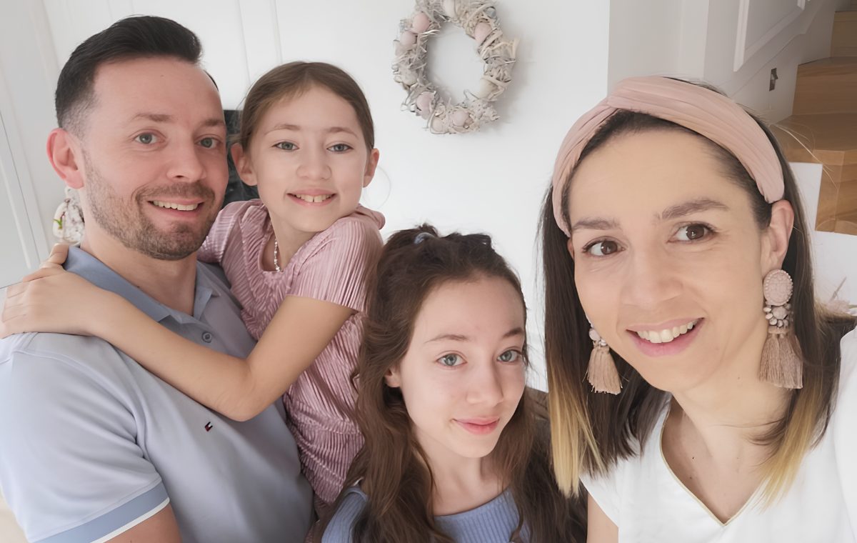 A smiling family of four, two parents with their two daughters, pose together for a warm close-up photo at home.