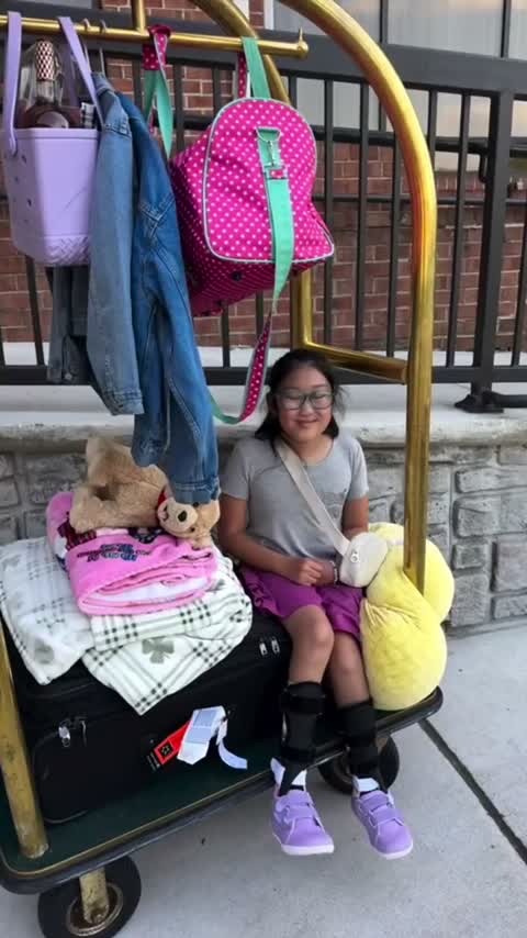 Una niña que lleva una tobillera ortopédica está sentada en un carrito de equipaje de hotel lleno de sus pertenencias.